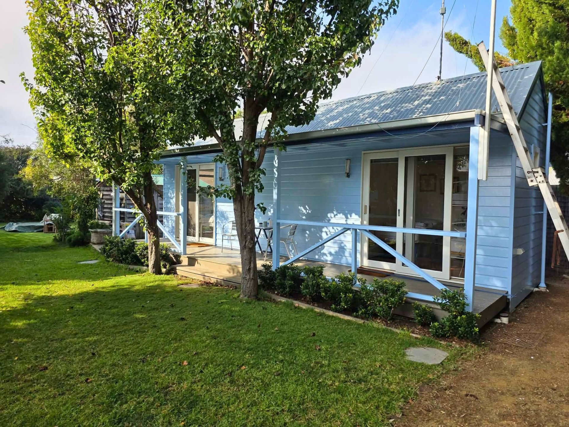 Light blue cottage with white veranda and solar roof panels surrounded by green lawn and large trees