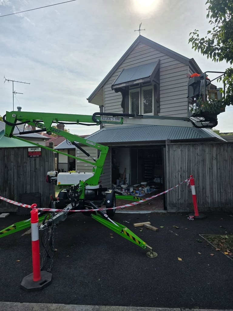 Green mobile elevated work platform parked outside a white church building with red and white safety barriers during exterior work
