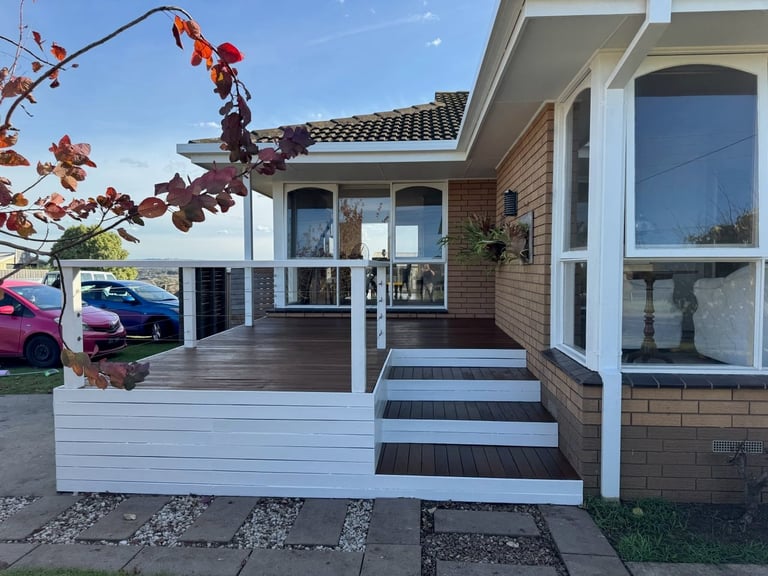 Modern brick and white home exterior with wooden deck, steps, large windows, parked car visible, and autumn foliage in front yard