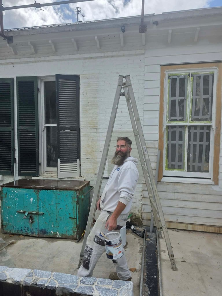 Man with beard standing beside a ladder against a white house exterior with windows and a turquoise storage box