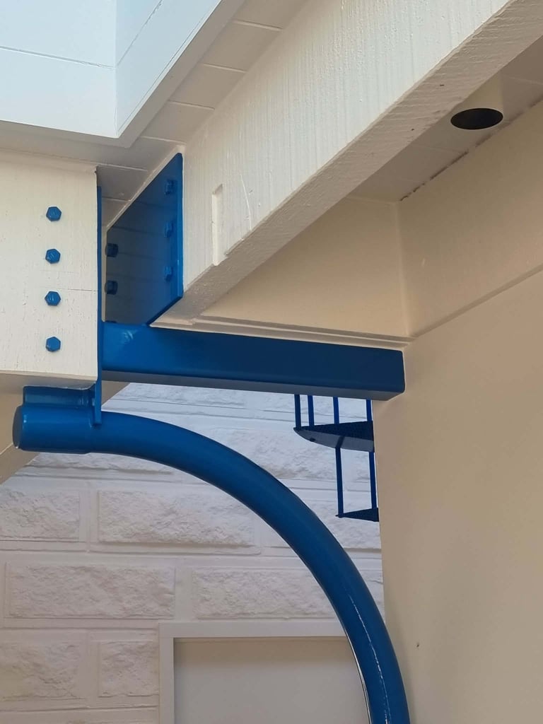 Blue curved metal pipe and structural support beam mounted on white wall with ceiling details and skylight above