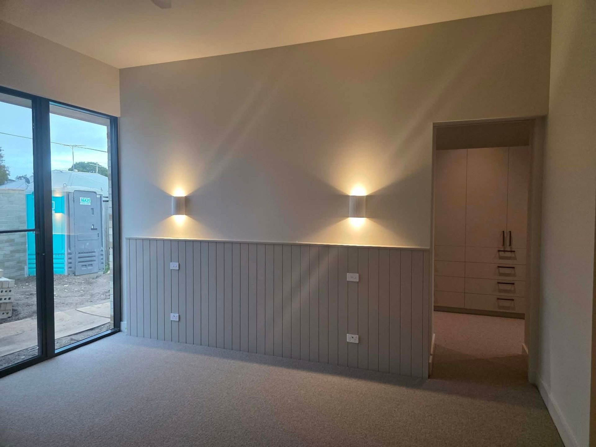Modern apartment interior with beige walls, gray horizontal paneling, uplighting, glass door, and doorway to adjoining room