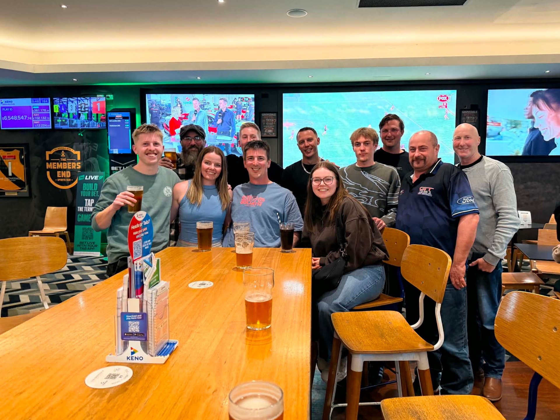 Group of about ten people posing together at a wooden table in a sports bar with multiple TV screens on the wall behind them