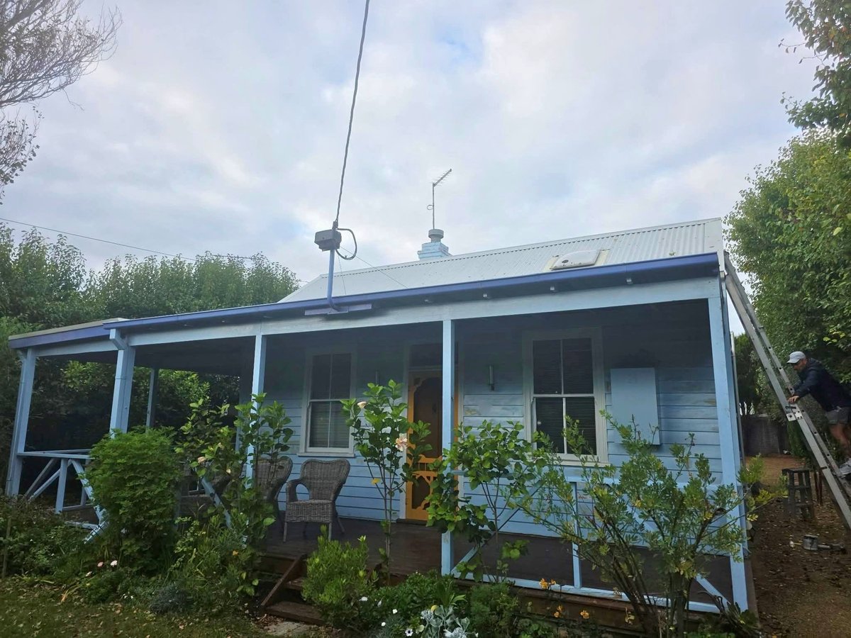 Small blue and white cottage with covered front porch, surrounded by green vegetation and trees