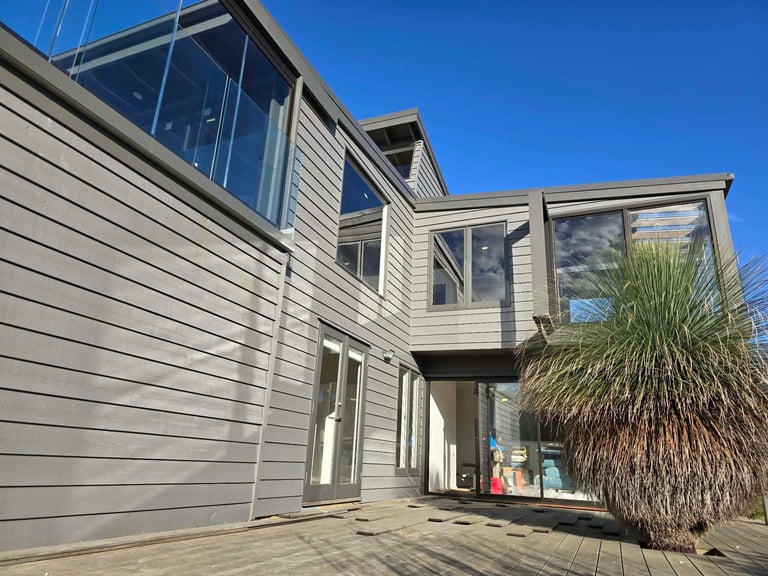 Modern house with light gray siding, large windows, wooden deck, and palm tree on clear blue sky day