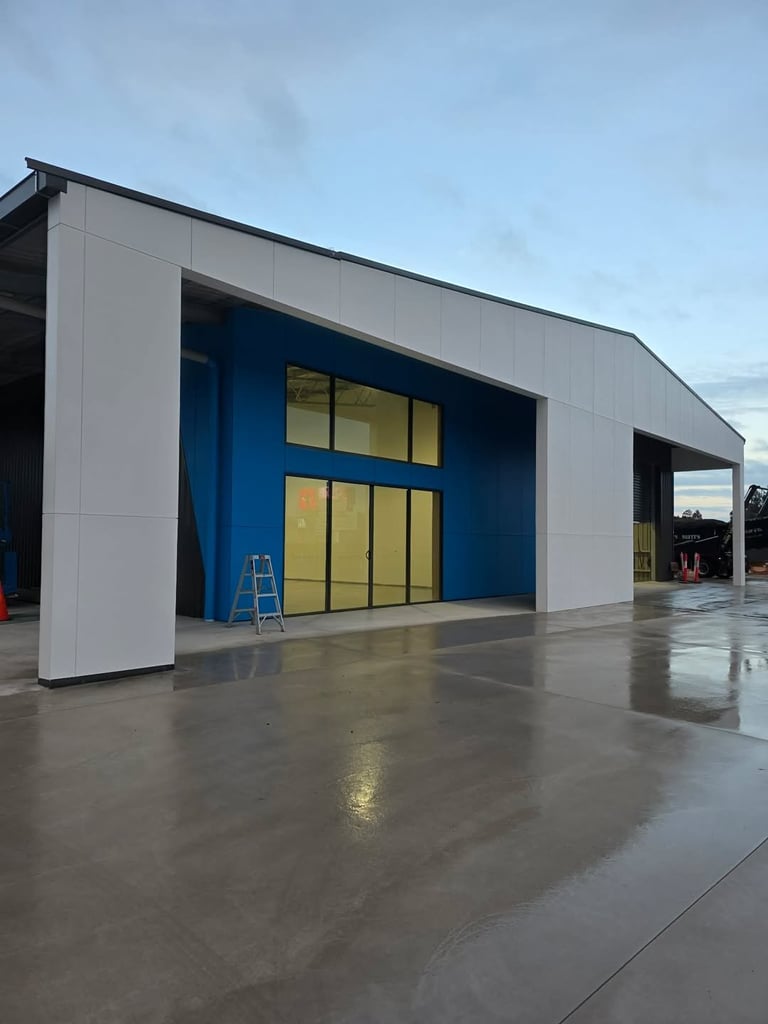 Modern industrial building with white panels and blue accent wall, large yellow-tinted glass doors, wet concrete foreground, overcast sky