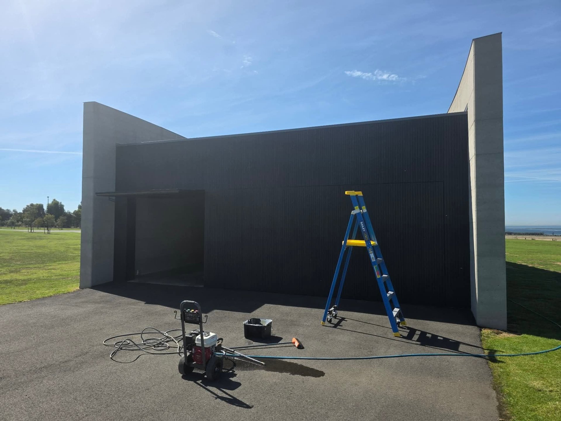 Modern black geometric building structure under construction with blue ladder and equipment on concrete pad in green field