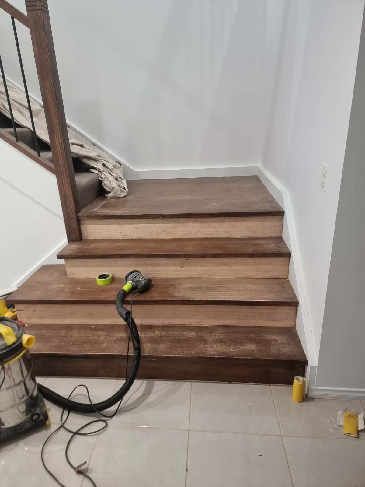 Wooden staircase under construction with tools and equipment scattered on floor, white walls and dark handrail visible