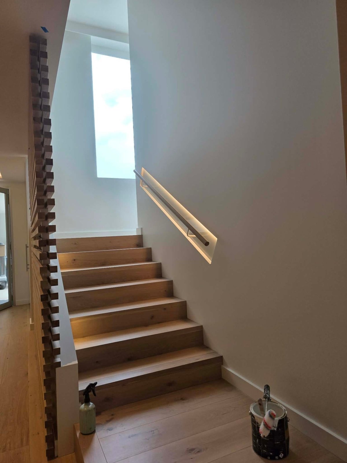 Modern wooden staircase with metal handrail leading upward toward skylight in bright, minimalist interior space