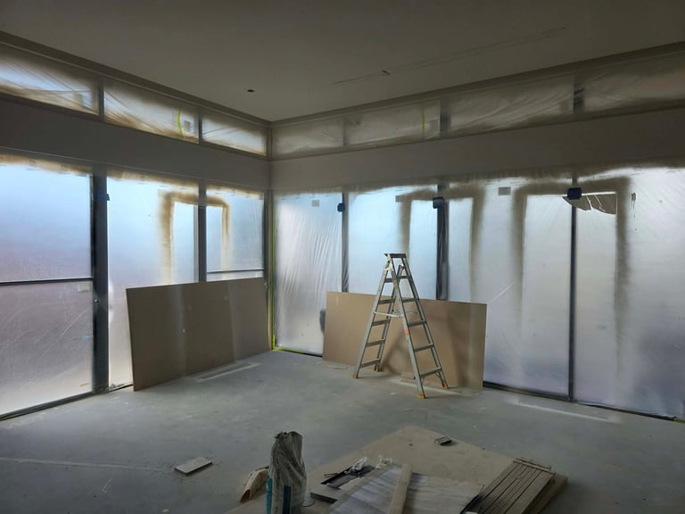 Construction site interior with plastic sheeting, ladder, and drywall panels in unfinished room with windows