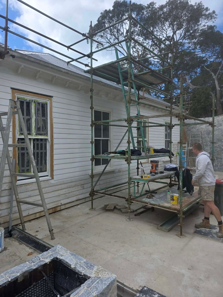 Worker on construction scaffold next to white weatherboard house undergoing renovation with trees in background