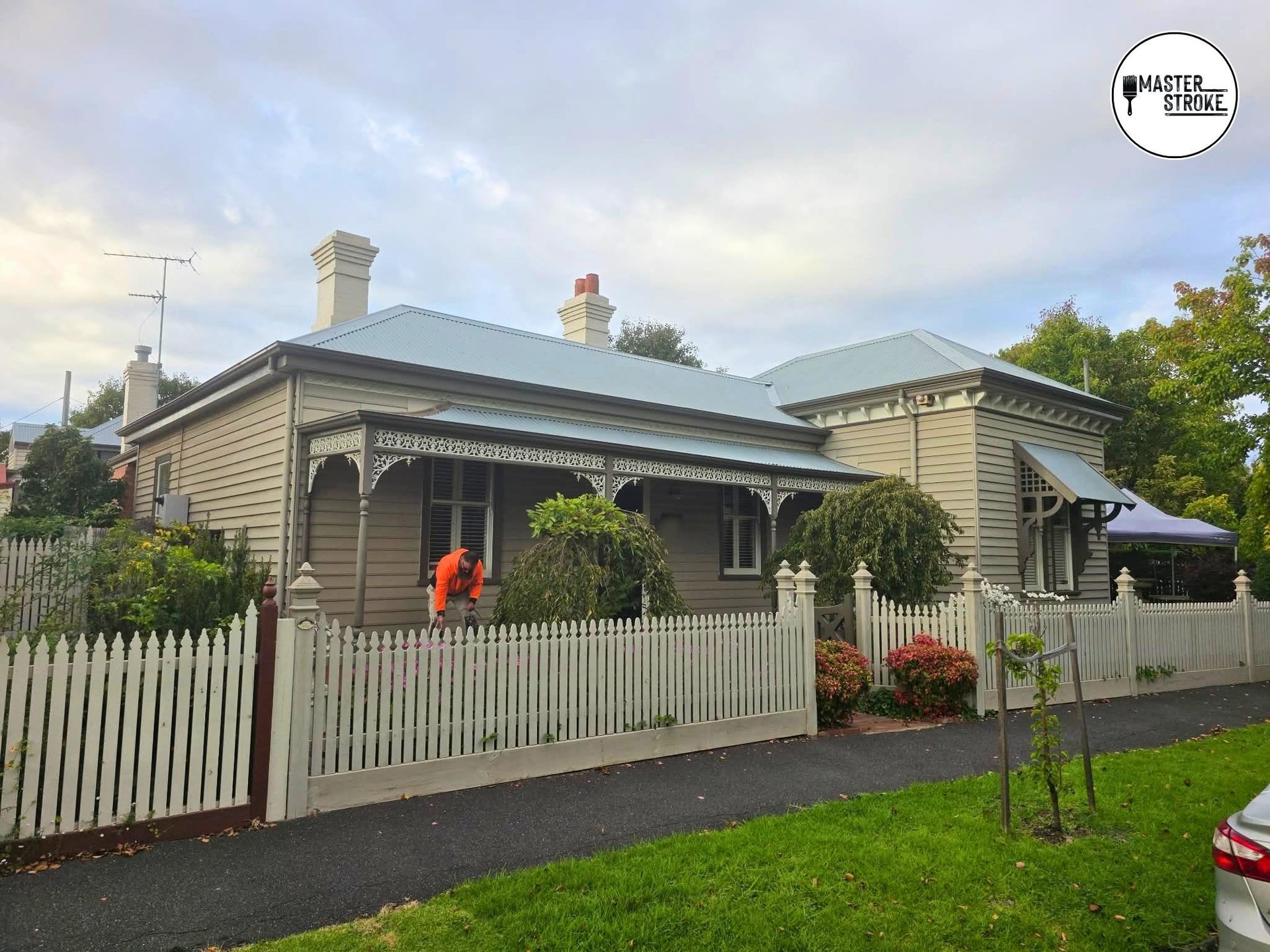 Cream weatherboard cottage with green roof, white picket fence, front garden, and brick chimneys on overcast day