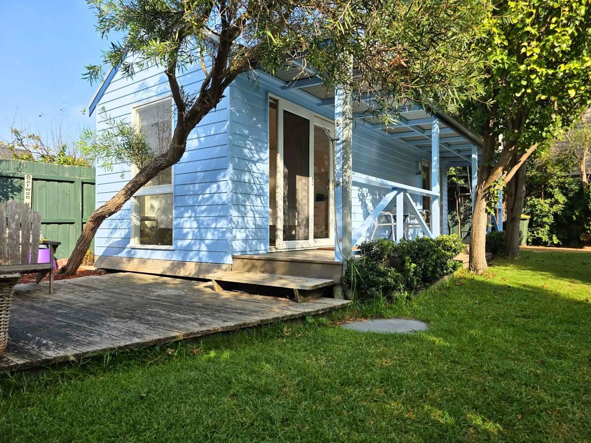 Light blue cottage with wooden deck and porch surrounded by mature shade trees and green lawn