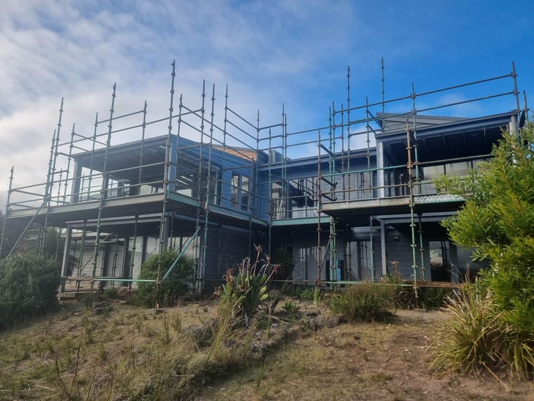 Multi-story house under construction with metal scaffolding and framework, surrounded by sandy terrain and green shrubs
