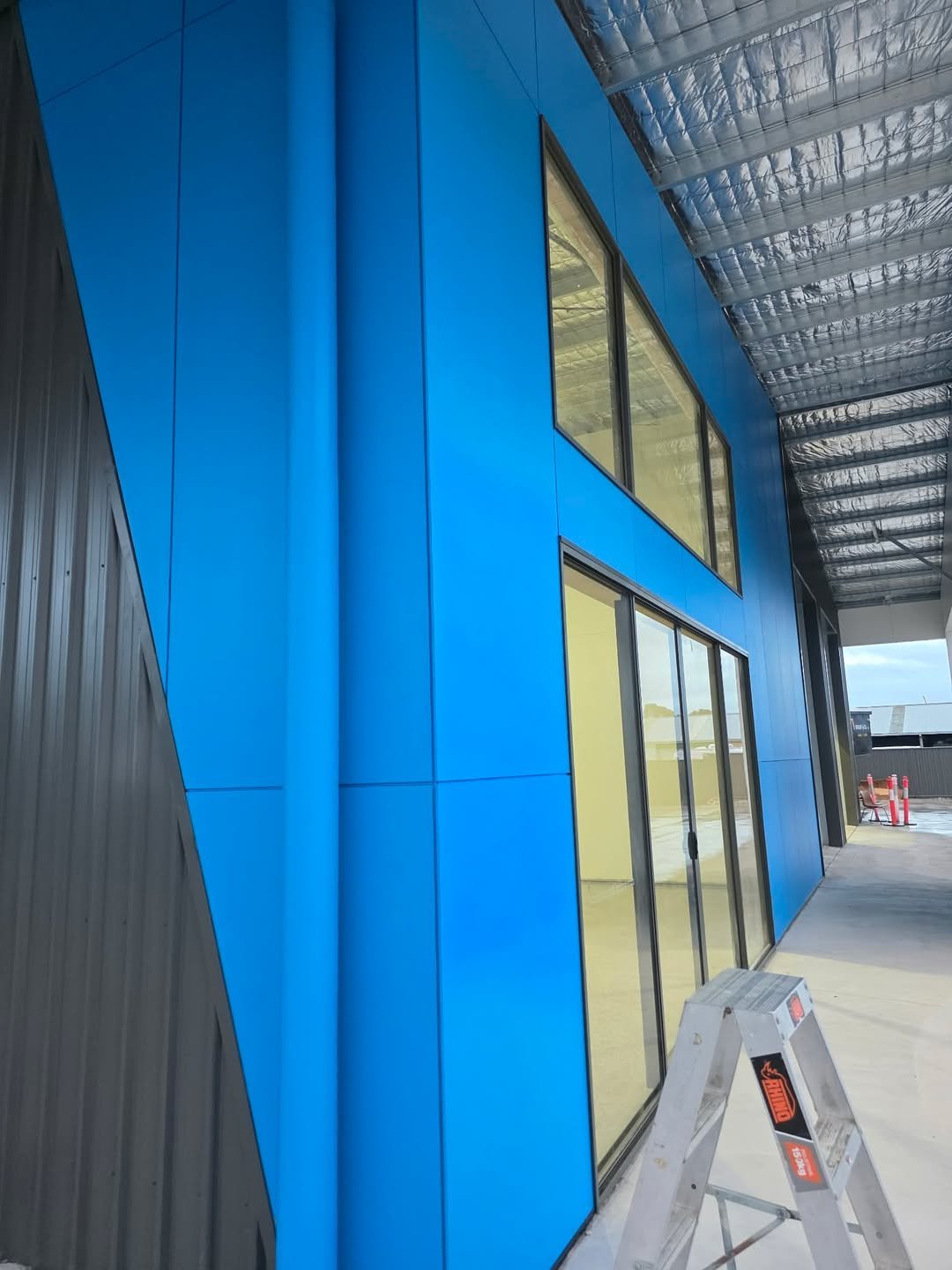 Modern blue metal building exterior with large windows and corrugated metal roof, ladder visible in foreground