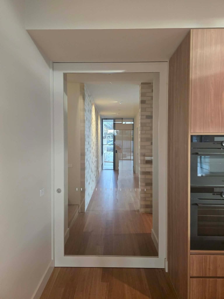 Modern kitchen with glass-paneled door opening to a bright hallway, featuring wooden flooring and stainless steel appliances on the right