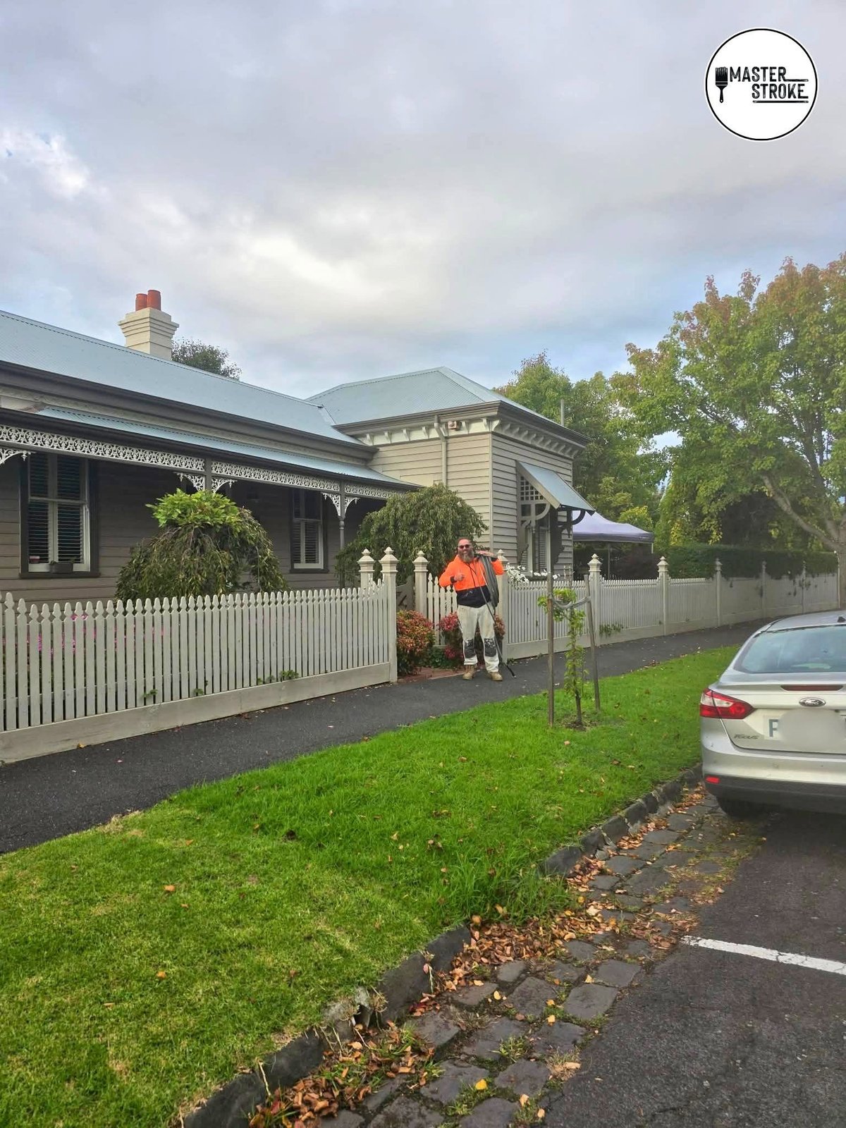 Victorian-style house with white picket fence, green lawn, and person in orange standing at the gate under cloudy sky