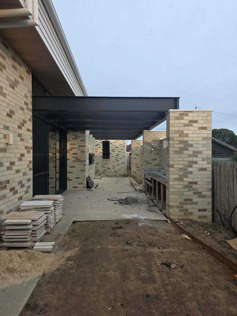 Under-construction residential patio area with tan brick walls, black pergola frame, and dirt ground during building renovation