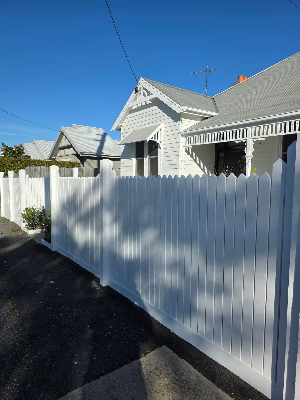 White wooden fence in front of a classic white house with a pitched roof and wraparound porch on a clear day