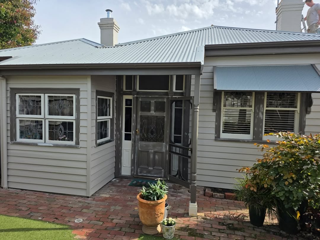 Gray cottage with metal roof, front entrance with screen door, brick patio, potted plants, and manicured garden