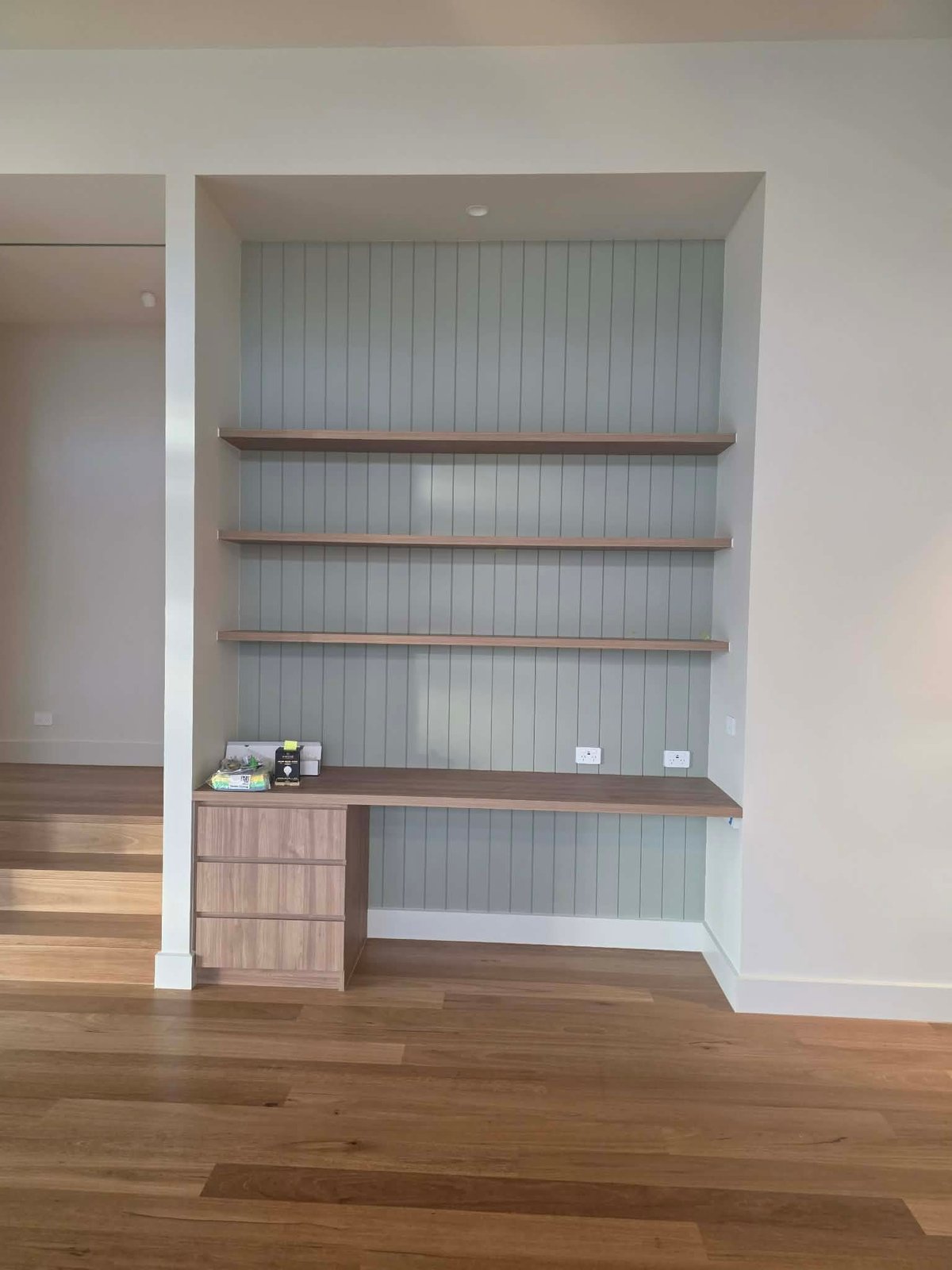 Built-in shelving unit with three wooden shelves, vertical paneled backdrop, wooden desk surface with cabinet below, and wood flooring