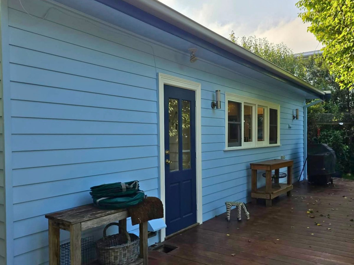 Light blue cottage exterior with dark blue door, wooden deck, and surrounding green foliage