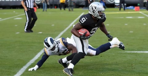 NFL player in dark uniform evading tackle while running with football, opponent diving behind