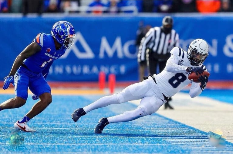 Football player in white jersey diving with ball while defender in blue jersey approaches during game