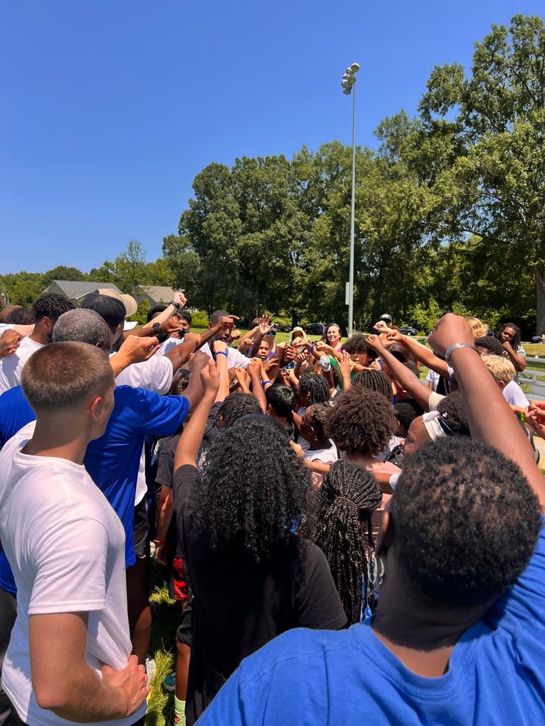 Large group of people huddled together outdoors with hands raised in the center, outdoor sports field setting with trees and tall light pole