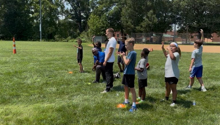 Group of young children participating in an outdoor sports training session on a grass field with a baseball diamond visible in the background