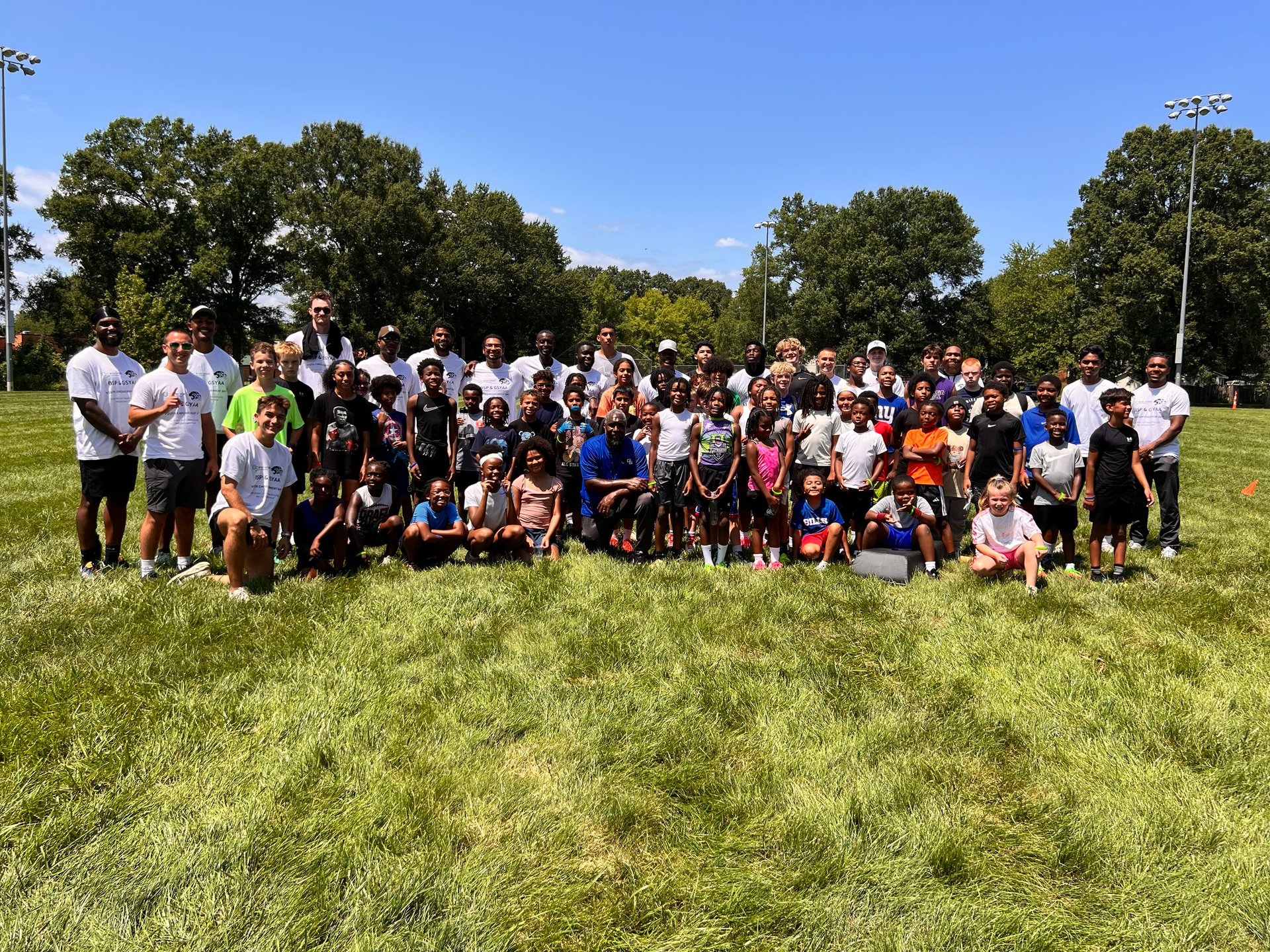 Large group of diverse children and adults standing together on a sunny green grass field with trees in the background
