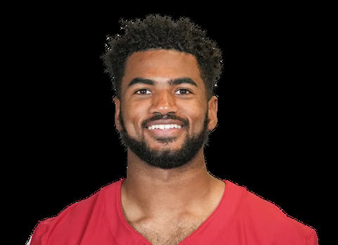 Headshot portrait of a smiling man with dark curly hair and beard wearing a red v-neck shirt against a black background