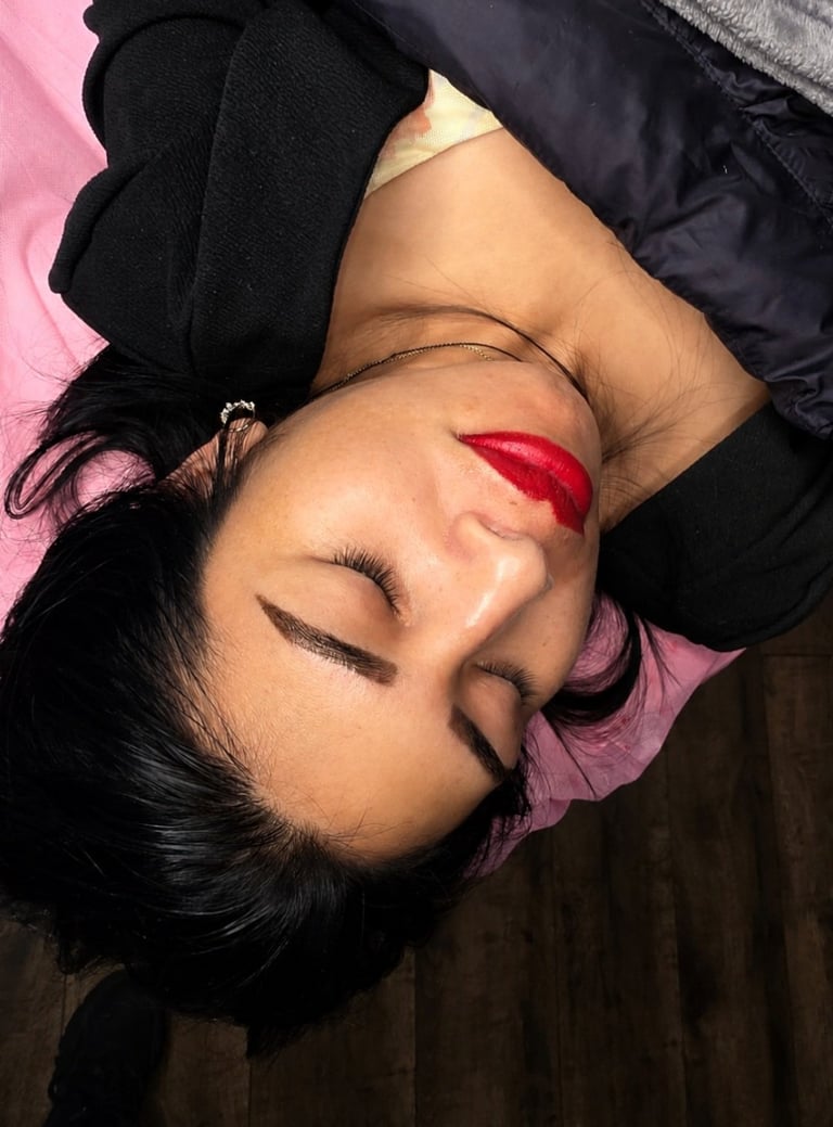 Person with long black hair lying on pink background, wearing dark clothing, with red lipstick and eyes closed