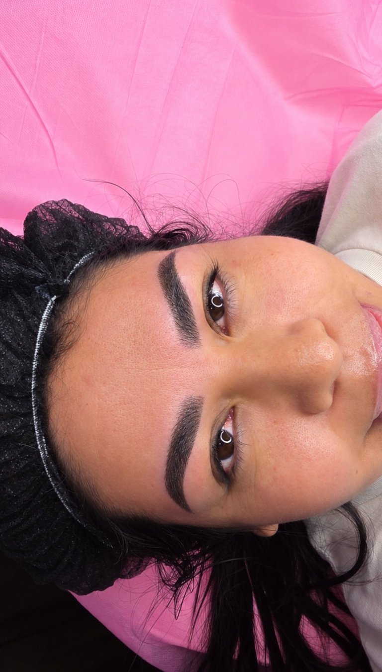 Close-up portrait of a person with defined eyebrows and makeup, wearing a pink tulle accessory against a pink background