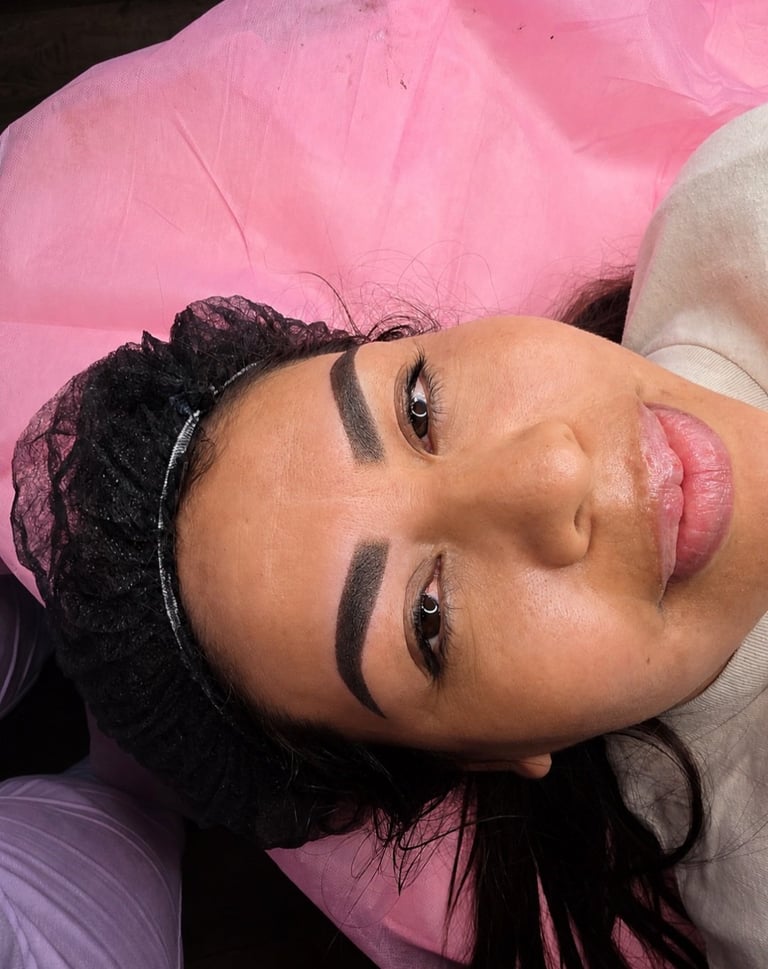 Person with dark braided hair wearing bold eyebrow makeup and pink lipstick, tilting head against pink fabric background