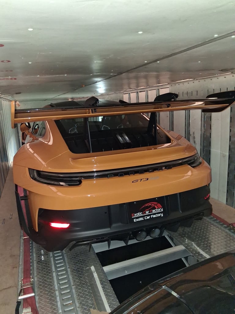 Orange Porsche GT3 parked on a vehicle lift in an underground garage with concrete ceiling and metal framework