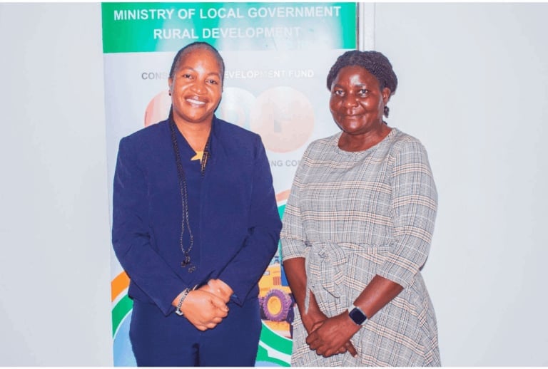 Two women posing together in front of a Ministry of Local Government and Rural Development banner at an official event