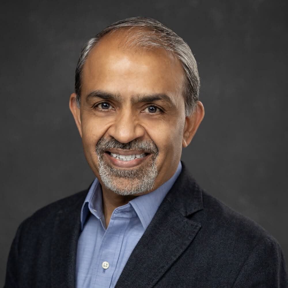 Professional headshot of a middle-aged man with gray hair and beard, wearing a dark blazer over a light blue shirt, smiling against a gray background