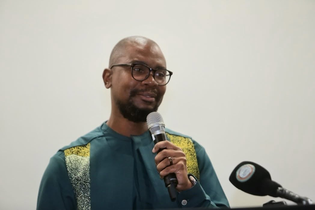 Man with glasses and beard wearing a teal and gold jacket speaking into a microphone