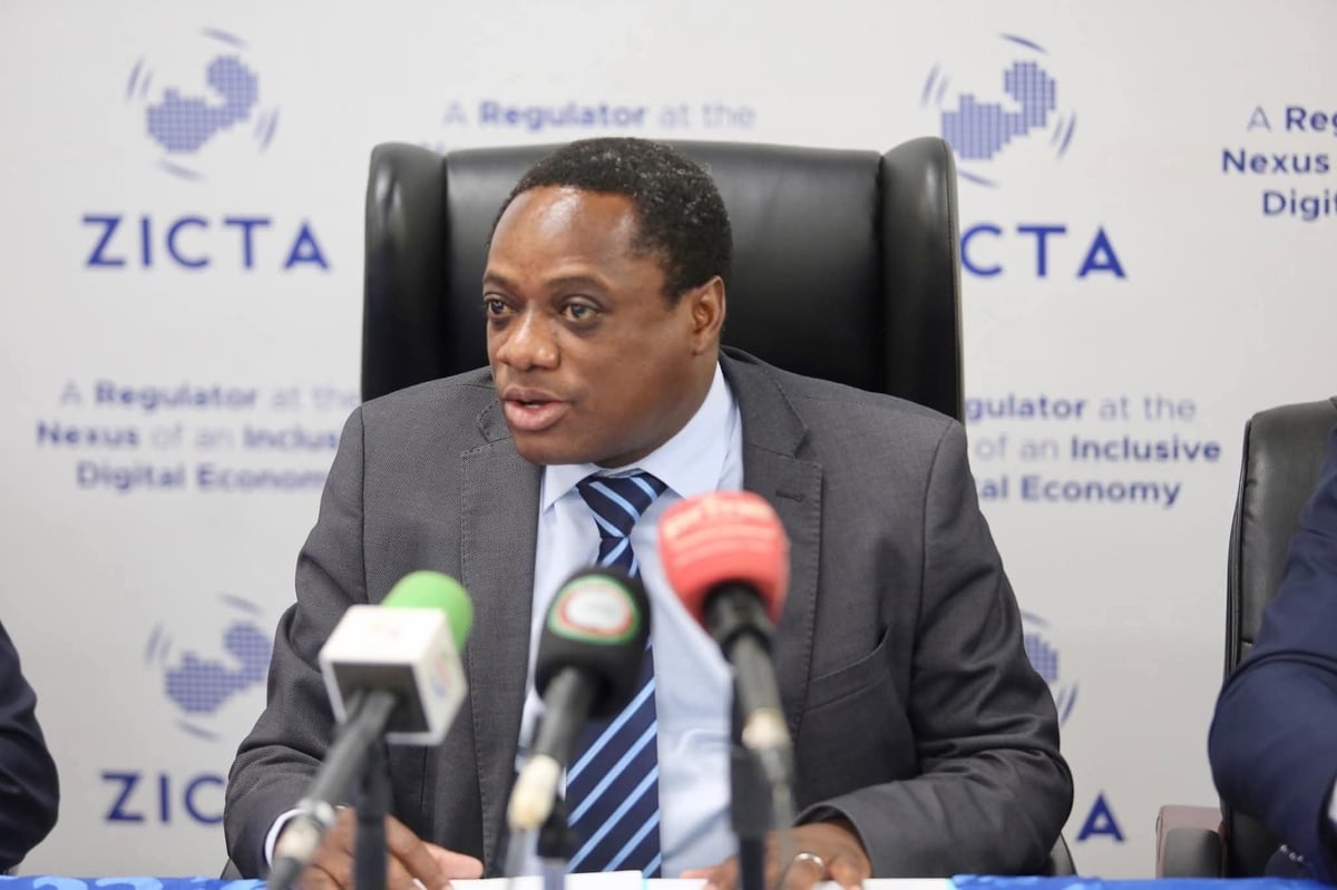 Man in gray suit speaking at microphones during press conference with ZICTA logo backdrop