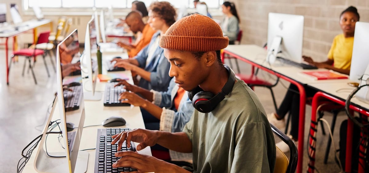 Man wearing orange beanie and headphones typing at computer in classroom with other students at desks in background