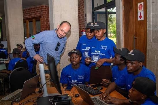 An instructor in light blue shirt demonstrates technology to a group of young students wearing matching blue shirts at computer workstations.