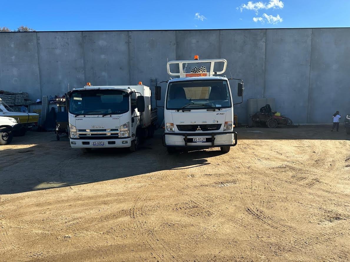 Two white commercial trucks parked on sandy ground against a gray concrete wall under blue sky