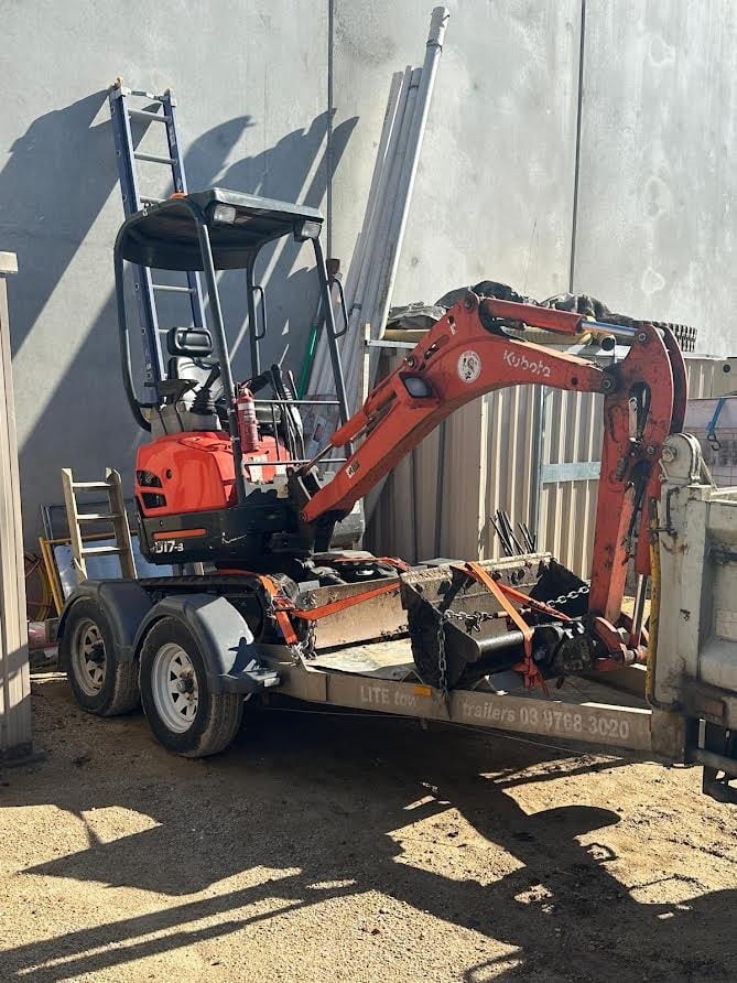 Red mini excavator mounted on a trailer parked against a metal building in bright sunlight