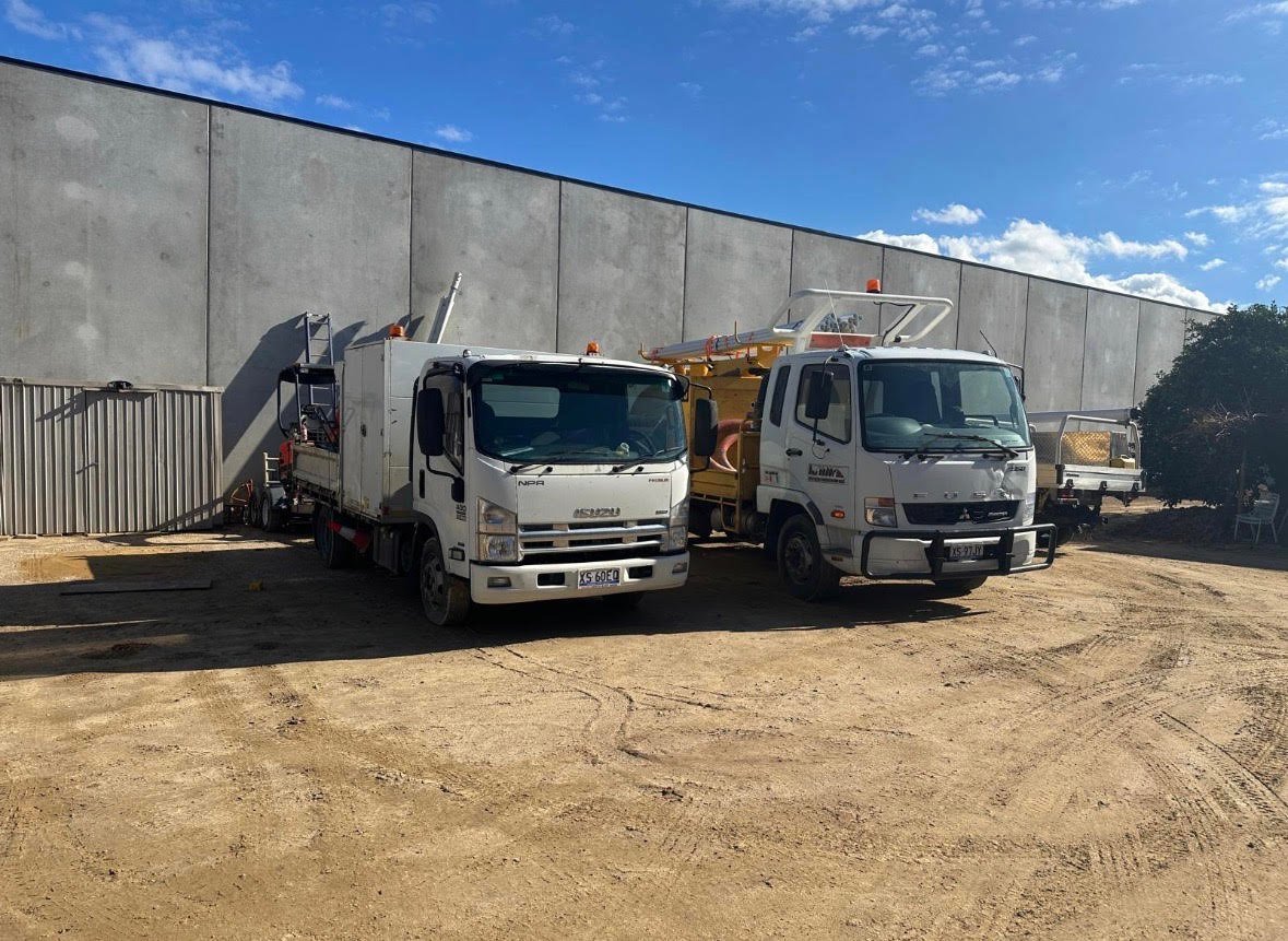 Two white industrial trucks parked at a construction site with a concrete warehouse building and blue sky in the background