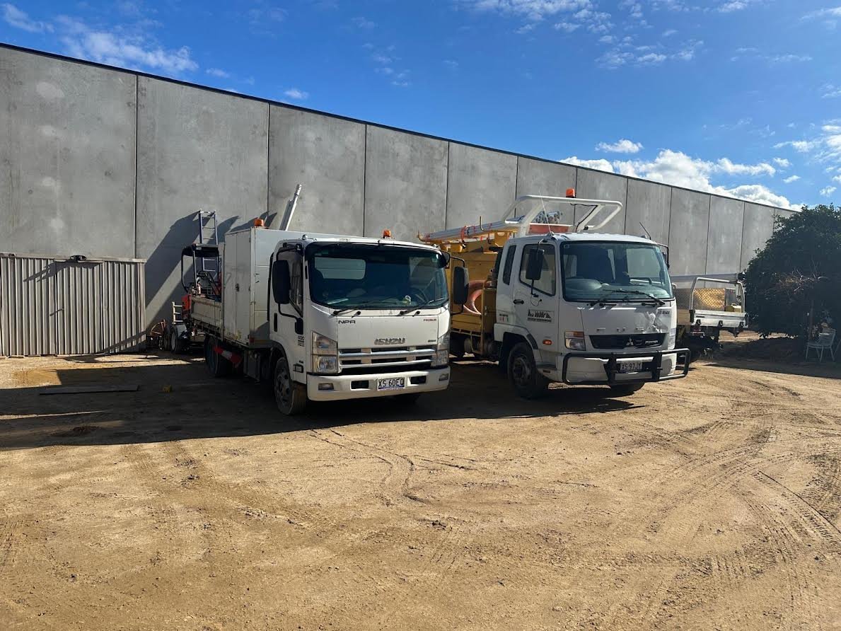 Two white industrial trucks parked on dirt ground in front of a large concrete warehouse building