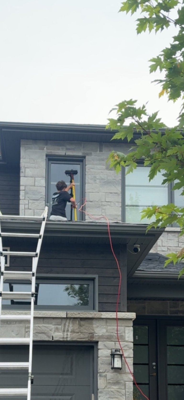 Worker on ladder using power drill on upper level of modern two-story house with stone and dark siding
