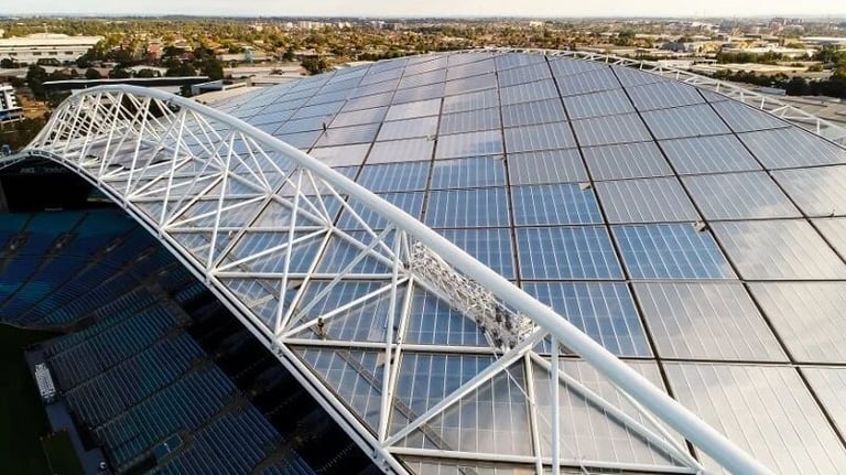 Aerial view of a stadium rooftop covered with blue solar panels and white structural framework supporting the installation
