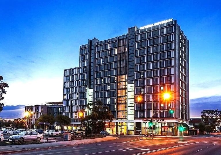 Modern multi-story residential building illuminated at dusk with street-level retail, surrounded by parked cars and trees