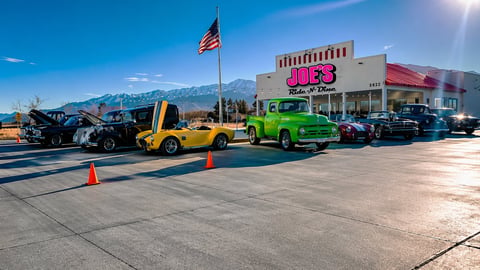 Colorful classic cars displayed in a parking lot with mountains and American flag in background at an auto show